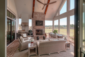 Tabby concrete floors with brick borders, v-groove ceilings, vaulted ceiling, brick fireplace, reclaimed timber mantle, outdoor kitchen. A screened porch with a vaulted ceiling, dining table, striped couch and two end tables, and a TV mounted over a brick fireplace with a reclaimed timber mantle. There is an outdoor kitchen in one corner.
