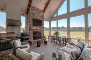 Tabby concrete floors with brick borders, v-groove ceilings, vaulted ceiling, brick fireplace, reclaimed timber mantle, outdoor kitchen. A screened porch with a vaulted ceiling, dining table, striped couch and two end tables, and a TV mounted over a brick fireplace with a reclaimed timber mantle. There is an outdoor kitchen in one corner.
