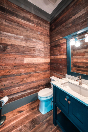 A powder room with rustic wood flooring and wall paneling. There is a small blue vanity with a marble countertop.