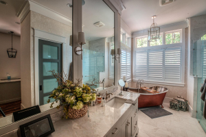 A lavish bathroom with marble floors and countertops. There is a gray vanity with mirrors in the foreground and a large copper soaking tub in the far corner.