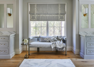 A large bathroom with a gray upholstered bench set between two gray vanities with marble countertops. The floor is medium brown hardwood with a marble inlay.