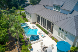 Overhead shot of the pool deck in back of a large white home with a dark metal roof.