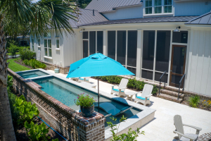 A backyard swimming pool with a spa on one end and a tanning ledge on the opposite end. There are lounge chairs and a blue umbrella on the tanning ledge.