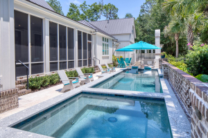 A backyard swimming pool with a spa on one end and a tanning ledge on the opposite end. There are lounge chairs and a blue umbrella on the tanning ledge.