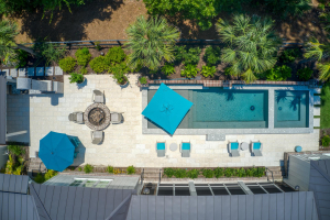 Overhead shot looking down backyard swimming pool with a spa on one end and a tanning ledge on the opposite end. There are lounge chairs and a blue umbrella on the tanning ledge. Nearby are a fire pit and a small umbrella-shaded round dining table and chairs.