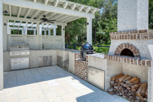 An outdoor kitchen with a Green Egg style cooker, a stainless steel gas grill, and a wood-fired oven. The counters and oven chimney are tabby stucco.