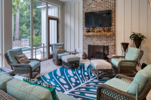 Brick fireplace, reclaimed timber mantle, tabby concrete floors, mahogany doors. 4 wicker chairs and a couch form a sitting area on a screened porch in front of a fireplace with a TV mounted above the reclaimed timber mantle.