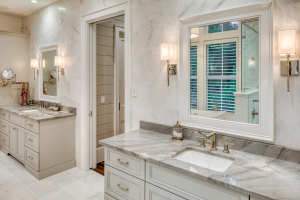 A master bathroom with light-colored marble walls and flooring. There are two light gray vanities with gray marble countertops and white undermount sinks.