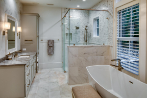 A master bathroom with light-colored marble walls and flooring. It features a white free-standing tub and marble shower opposite two light gray marble top vanities with white undermount sinks.
