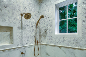 A shower with small gray marble tiles of varied shades laid in a herringbone pattern. The showerhead is gold.