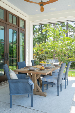 Outdoor dining area, tabby shell concrete floors, haint blue v-groove ceiling, mahogany doors, motorized retractable screens. A brown wooden table with blue chairs sits on the back porch of a large home.
