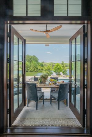 Outdoor dining area, tabby shell concrete floors, haint blue v-groove ceiling, mahogany doors, motorized retractable screens. Brown mahogany double doors open onto a back porch with a wooden dining table and blue chairs overlooking a pool with lake views.