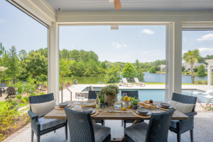 Outdoor dining area, tabby shell concrete floors, haint blue v-groove ceiling, motorized retractable screens. A wooden dining table with blue chairs overlooking a pool with lake views.