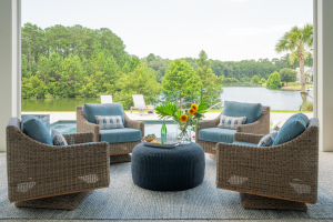 Outdoor sitting area, tabby shell concrete floors, motorized retractable screens. 4 brown wicker chairs with blue cushions are arranged in a circle around a small round blue table on a porch overlooking a pool with lake views.