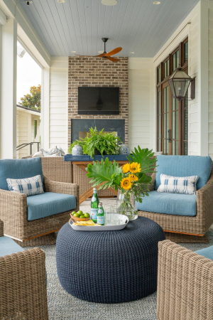 Outdoor sitting area, tabby shell concrete floors, haint blue v-groove ceiling, motorized retractable screens, outdoor fireplace, mahogany doors, Bevolo gas lanterns. 4 brown wicker chairs with blue cushions are arranged in a circle around a small round blue table on a porch with a TV mounted above an outdoor fireplace in the background.