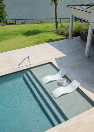 Two curvy white lounge chairs sit partially submerged in shallow end of swimming pool overlooking a lake.