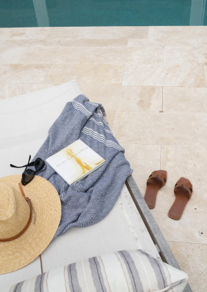 A woman's straw hat, sunglasses, towel, and book sit atop a lounge chair next to a pool's edge. There is a pair of brown sandals on the pool deck next to the chair.