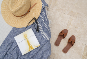 A woman's straw hat, sunglasses, towel, and book sit atop a lounge chair. There is a pair of brown sandals on the pool deck next to the chair.