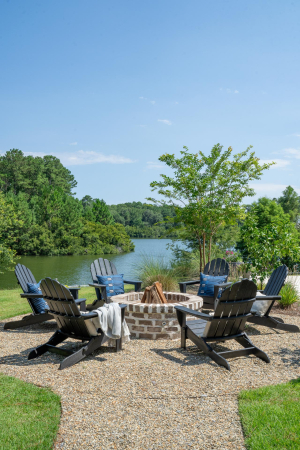 Six blue adirondack chairs are arranged in a circle around a circular brick fire pit overlooking a lake.
