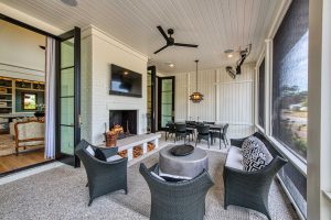 Tabby concrete floors, custom stone hearth with wood storage, painted brick fireplace, ceiling-mounted porch heaters, vertical shiplap siding, and a v-groove ceiling. A large screened porch with a TV mounted on a white painted brick fireplace, siting area, and a dining table.