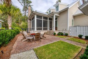 A large, round above-ground metal fire pit is surrounded by 6 wooden adirondack chairs. The fire pit area sits behind a home with beige siding and white trim, and it is defined by herringbone pattern brick hardscaping.