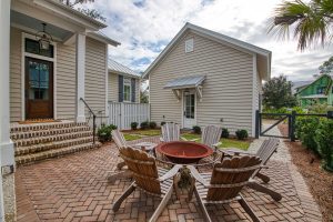 A large, round above-ground metal fire pit is surrounded by 6 wooden adirondack chairs. The fire pit area sits behind a home with beige siding and white trim, and it is defined by herringbone pattern brick hardscaping.