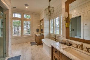 A large master bathroom with neutral tone shiplap walls, a large white pedestal soaking tub set between two custom rustic oak vanities with light colored marble tops, and light colored marble flooring.
