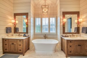 A large master bathroom with neutral tone shiplap walls, a large white pedestal soaking tub set between two custom rustic oak vanities with light colored marble tops, and light colored marble flooring.