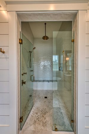 A large walk-in shower with a frameless glass door enclosure, built-in bench tiled seat, floor-to-ceiling light gray subway tile, and gray/white herringbone patter marble tile flooring. The shower features a bronze handheld shower and a bronze ceiling-mounted rain shower head.