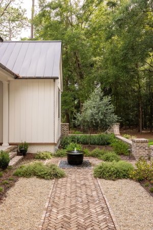 Tranquil garden with brick walkway, central fountain, and lush greenery beside a white board and batten home.