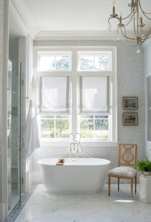 Freestanding soaking tub under large windows with Roman shades in soft neutral bathroom.