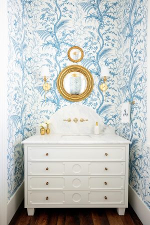 Elegant powder room with blue and white botanical wallpaper, marble vanity, and round gold mirror.
